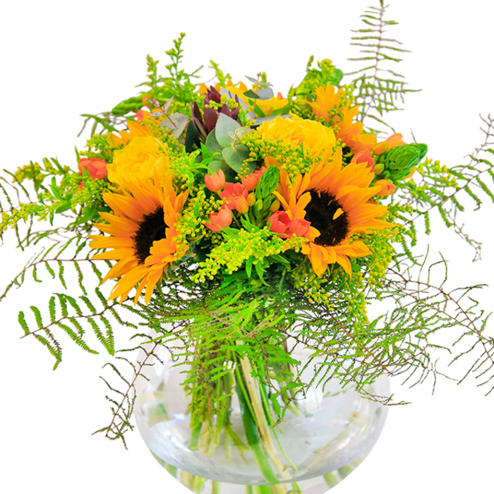 The initial impression of this bouquet is a wave of radiant, sun-inspired cheerfulness, reminiscent of summer afternoons by the River Lee. Towering sunflowers with golden petals and mocha centres anchor the design, exuding confidence and positivity. Intertwined among them, tight spirals of yellow garden roses bloom in lush abundance, their velvety petals catching and diffusing sunlight in gentle waves. Throughout, clusters of glossy red Hypericum berries provide fiery accents, a nod to the vibrancy found in Stratford's creative quarters and bustling high streets. Lush greenery spills out in every direction-feathery fern fronds, fresh solidago blossoms, and silvery eucalyptus leaves crafting texture and movement within the arrangement. Every stem is displayed in a perfectly clear, round vase, the water amplifying the vibrancy of the stems while giving the bouquet a sense of airy openness. Illuminated with soft, natural light, the arrangement shimmers, inviting touch and evoking scents of open fields and new beginnings. This hand-tied composition from Flowers Stratford suits joyful milestones-birthdays, anniversaries, or community gatherings-and brings a distinctive East London touch to every celebration, bridging nature's beauty with the heart of Stratford.